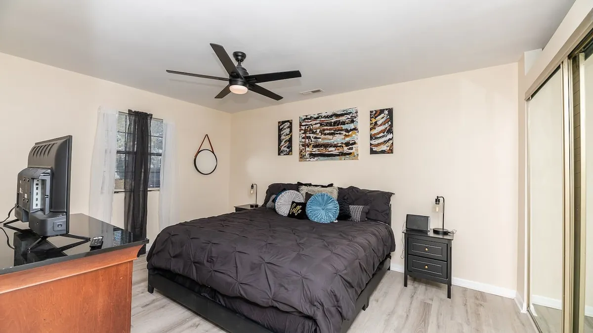 A large bed with sleek black bedding stands as the centerpiece, complemented by abstract wall art and a nightstand with a stylish lamp, ideal for creating customizable ambience. The overall setting is uncluttered and tranquil, emphasizing peaceful rest and modern comfort.