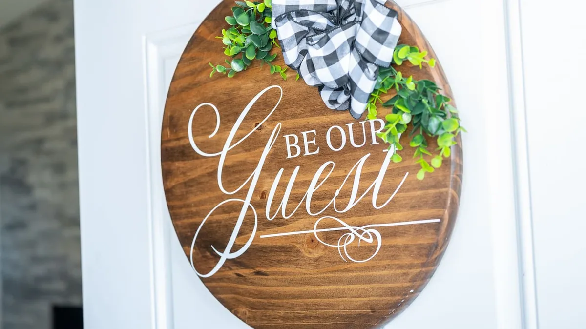 A bright and welcoming entryway adorned with a wooden sign, lush greenery, and a decorative bow conveys a sense of warmth and hospitality. The design cues suggest a space where guests are greeted with charm and care. The setup hints at a distinct foyer area that sets the tone for privacy and comfort upon entering the home.