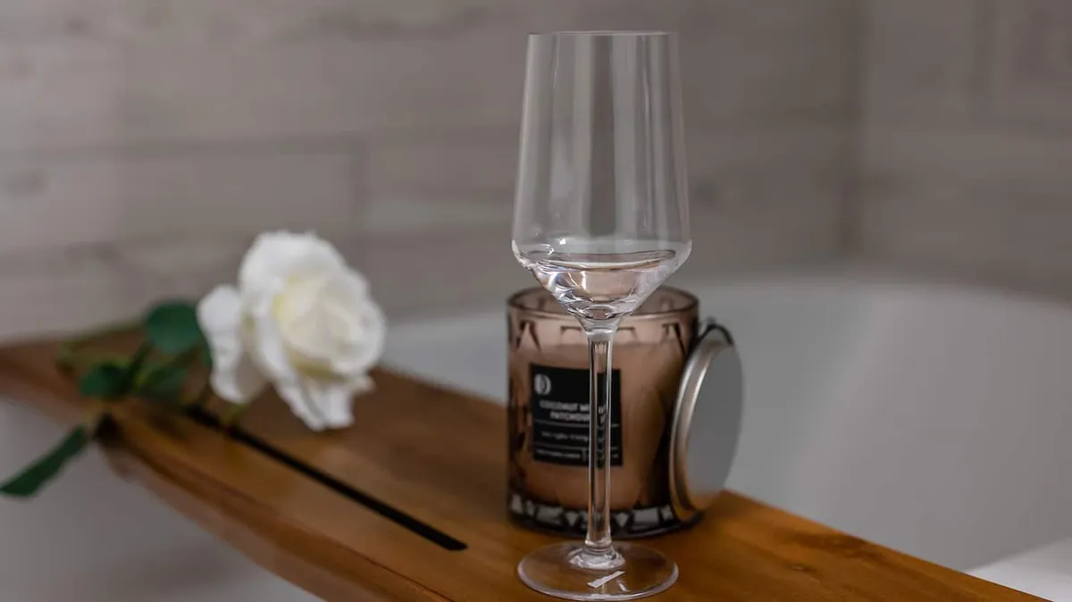 A serene bathroom scene with a wooden bath tray holding a wine glass and a white rose, complemented by a candle, suggesting a spa-like, tranquil environment ideal for unwinding after a busy day. The mood is calming, inviting moments of personal relaxation.