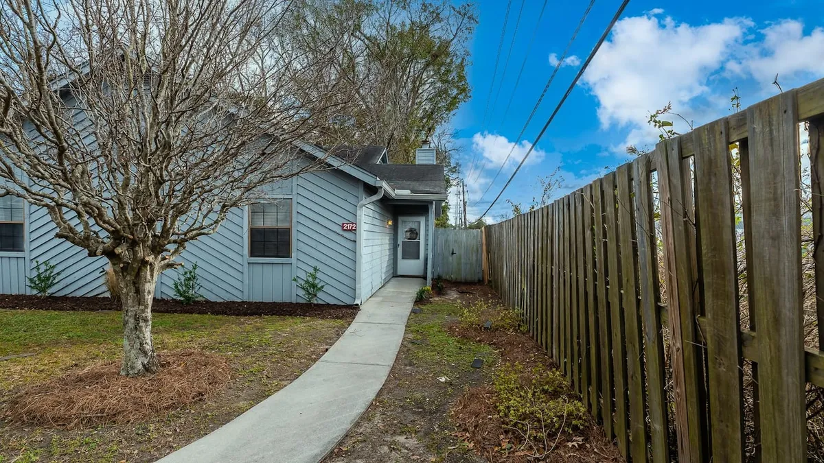 This image features the exterior of the townhouse, with a welcoming pathway leading up to the front door. A mature tree and a wooden fence frame the entrance, creating an inviting and peaceful atmosphere that hints at the home’s connection to the outside world.