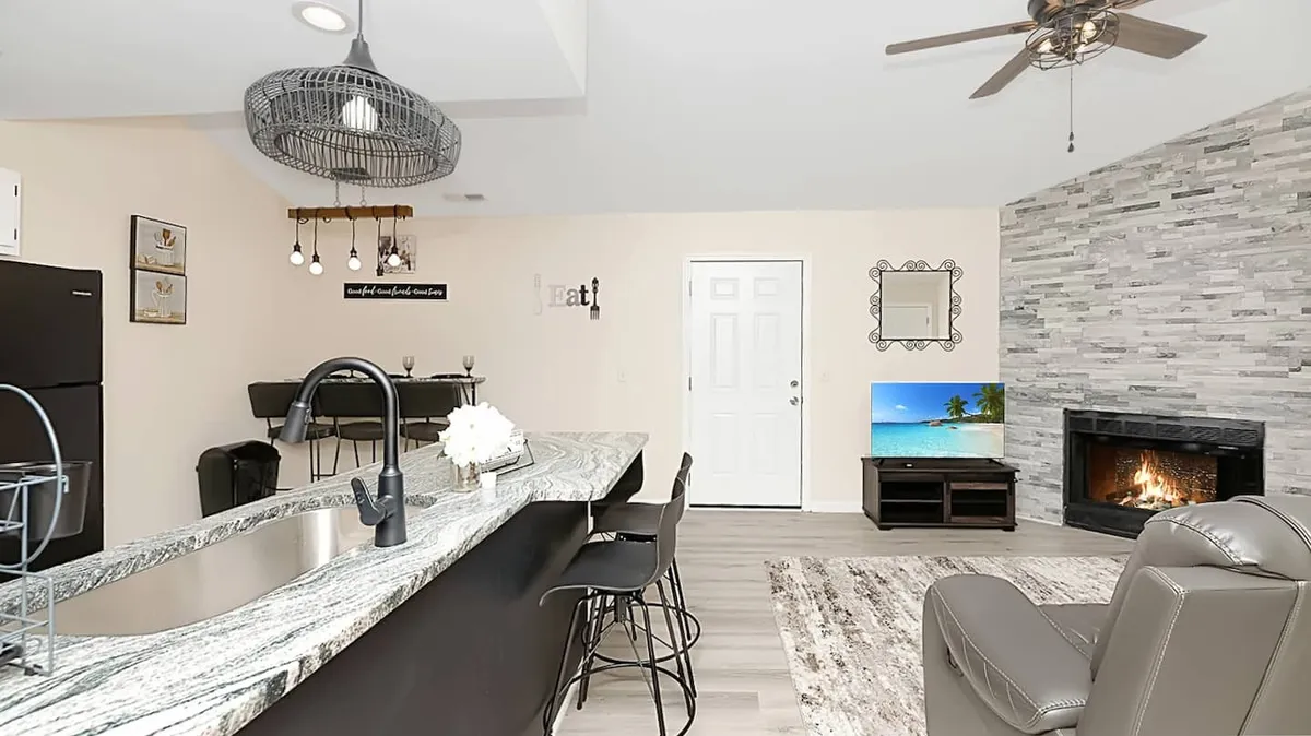A spacious open-plan living area featuring a kitchen island with bar stools, a fireplace, a television displaying a beach scene, and decorative wall art. The space is filled with natural light and has a welcoming, communal atmosphere perfect for gathering.