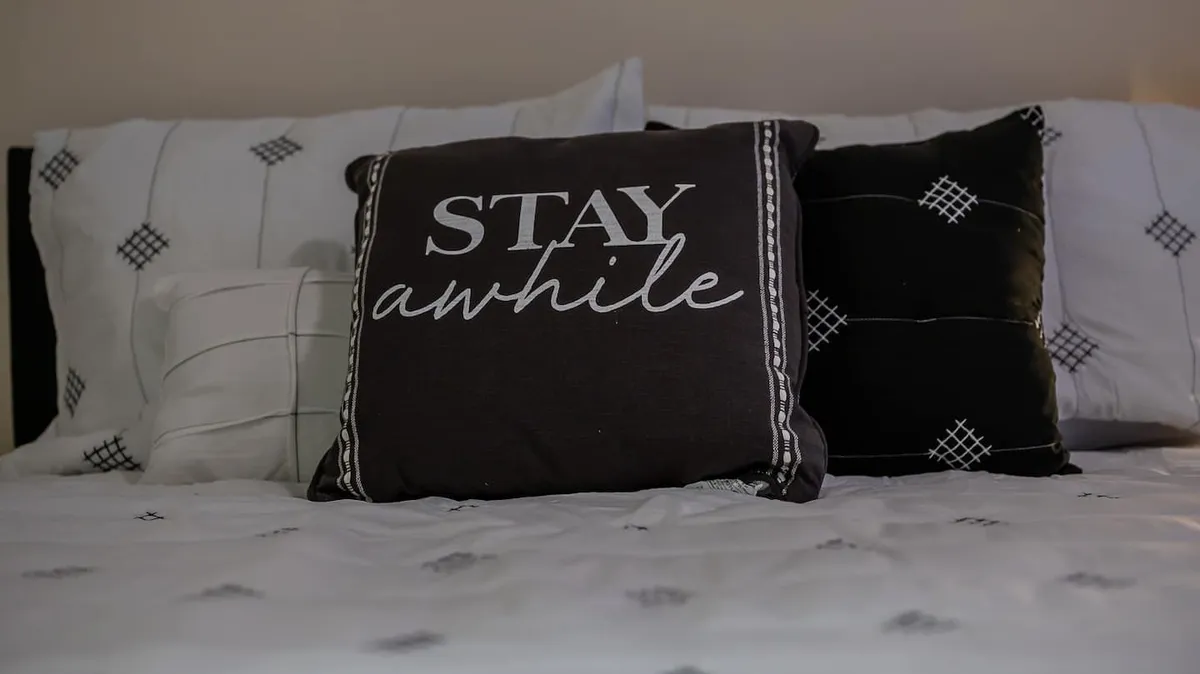 A close-up view of a bed adorned with a large decorative pillow reading 'STAY awhile', surrounded by smaller patterned pillows and plush bed linens. The soft textures and thoughtful arrangement create an inviting and restful atmosphere, ideal for relaxing, reading, or napping.