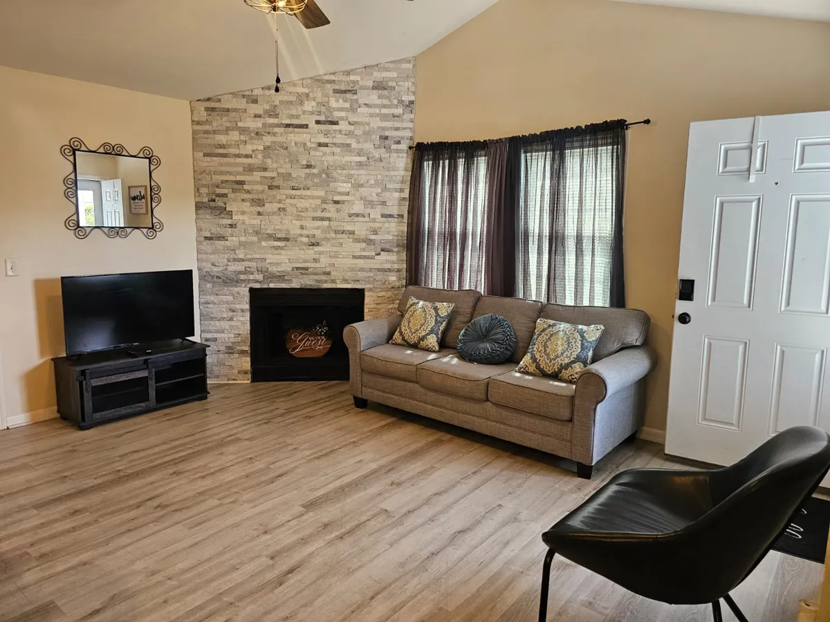 A cozy living room scene with a plush sofa, a stonewall fireplace, a television, a decorative mirror, and windows allowing in natural light. The setting is perfect for relaxing, watching TV, or socializing with family and friends.
