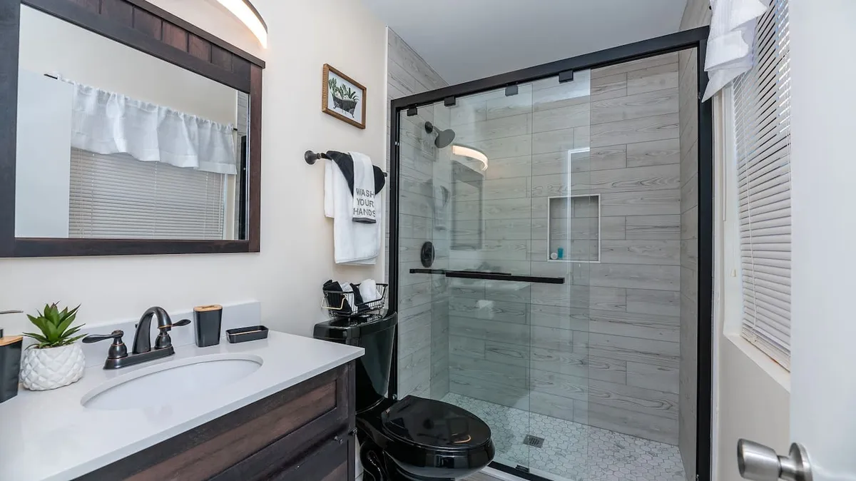 The image showcases a bright, modern bathroom with a sleek vanity, a glass-enclosed shower, a large mirror, plush towels, and a decorative plant, all set against a stylish, monochrome design. The space feels clean, inviting, and thoughtfully appointed.
