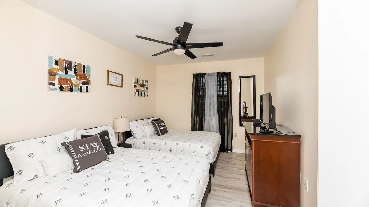 A spacious bedroom with two beds, decorative wall art, a ceiling fan, and a TV. Morning light filters in, creating a cozy yet sociable space ideal for friends or family to relax together while still enjoying privacy.
