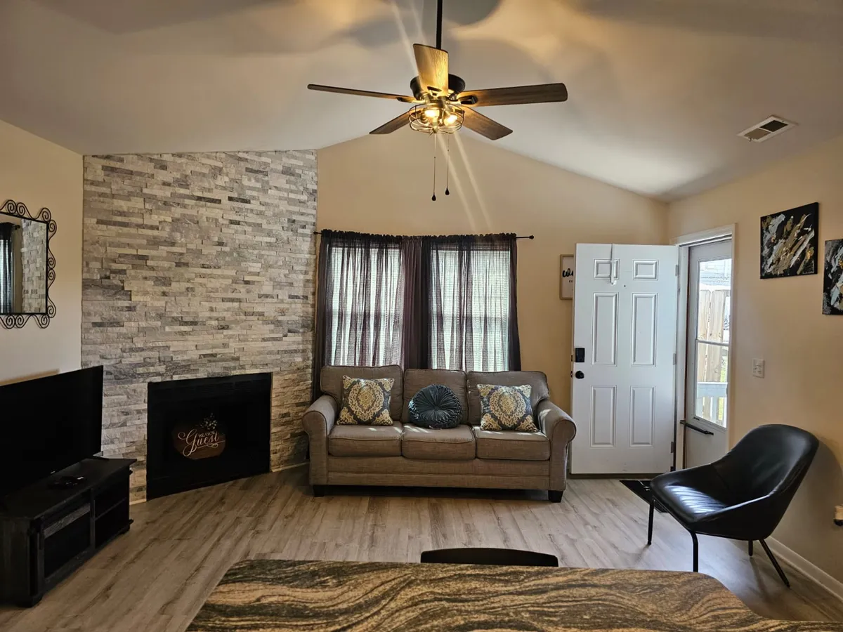 A light-filled family room with a prominent stone fireplace, a plush sofa decorated with stylish cushions, large windows that let in natural sunlight, and a modern black accent chair. The space is airy and inviting, ideal for both relaxing and entertaining guests.